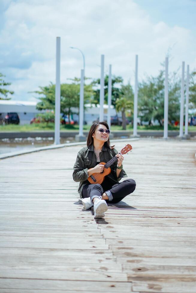 spelen ukulele van jong mooi Aziatisch vrouw vervelend jasje en zwart jeans poseren buitenshuis foto