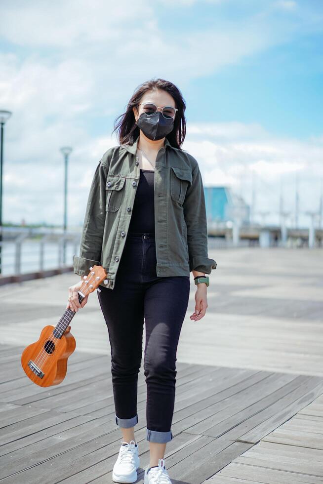 spelen ukulele van jong mooi Aziatisch vrouw vervelend jasje en zwart jeans poseren buitenshuis foto