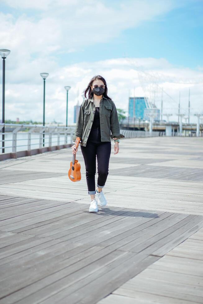 spelen ukulele van jong mooi Aziatisch vrouw vervelend jasje en zwart jeans poseren buitenshuis foto