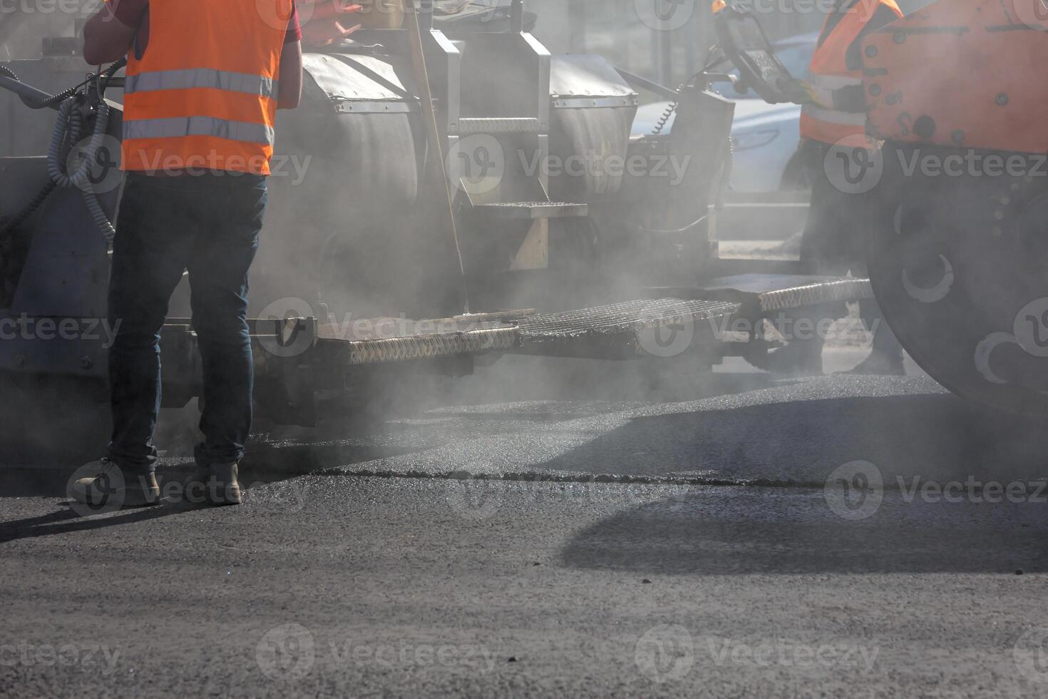 mannen werken met asfalteren bestratingsafwerkmachine machine gedurende weg straat repareren werken Bij dag licht met rook en stoom- in de lucht foto