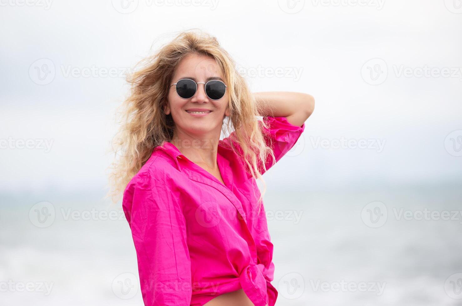 een mooi vrouw in zonnebril en een roze overhemd poses tegen de backdrop van de zee. foto