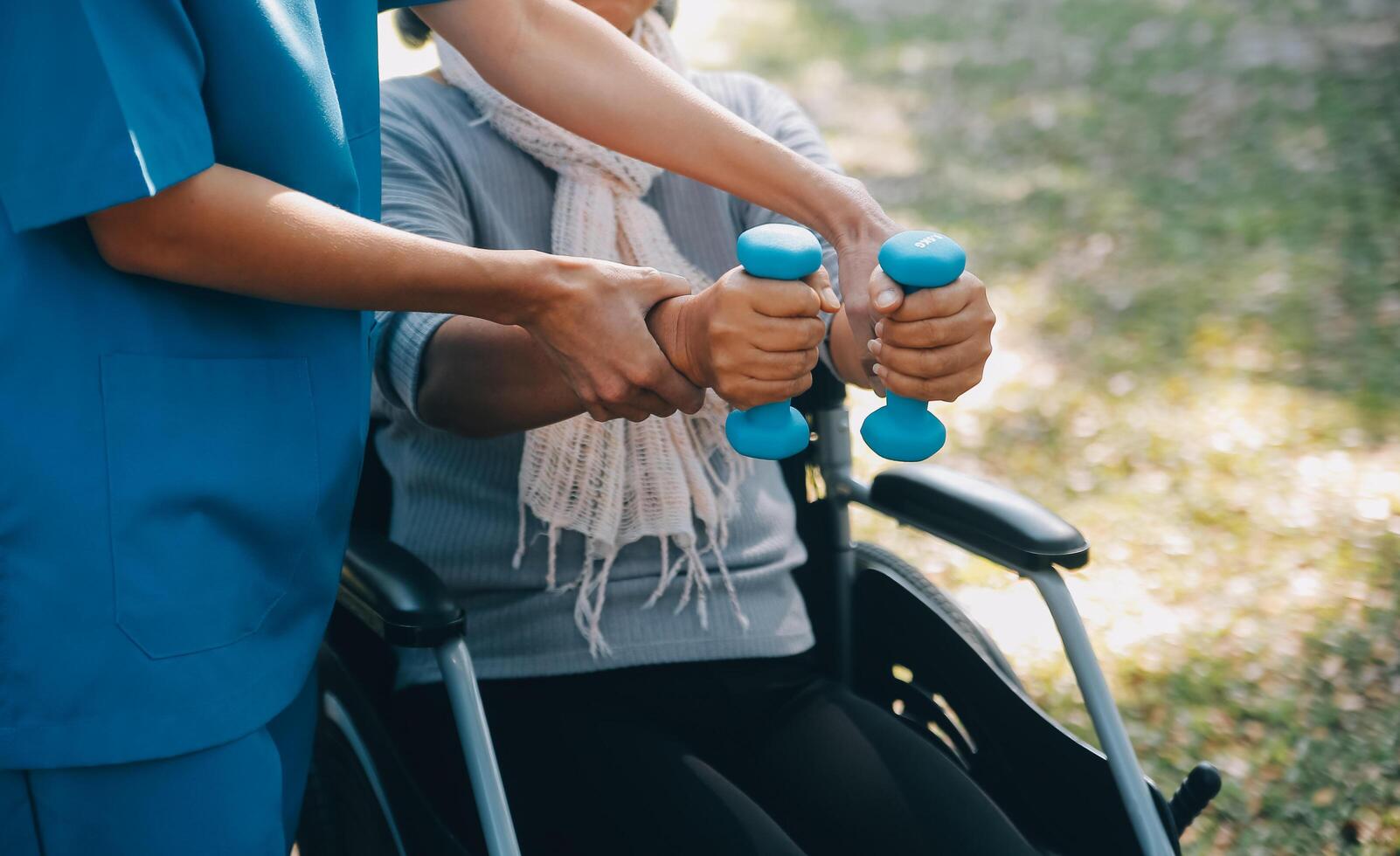 jong verpleegster of fysiotherapeut in scrubs helpen een gelukkig gepensioneerd oud vrouw Doen geschiktheid opdrachten met licht gewicht halters Bij huis. concept van fysiotherapie voor senioren foto