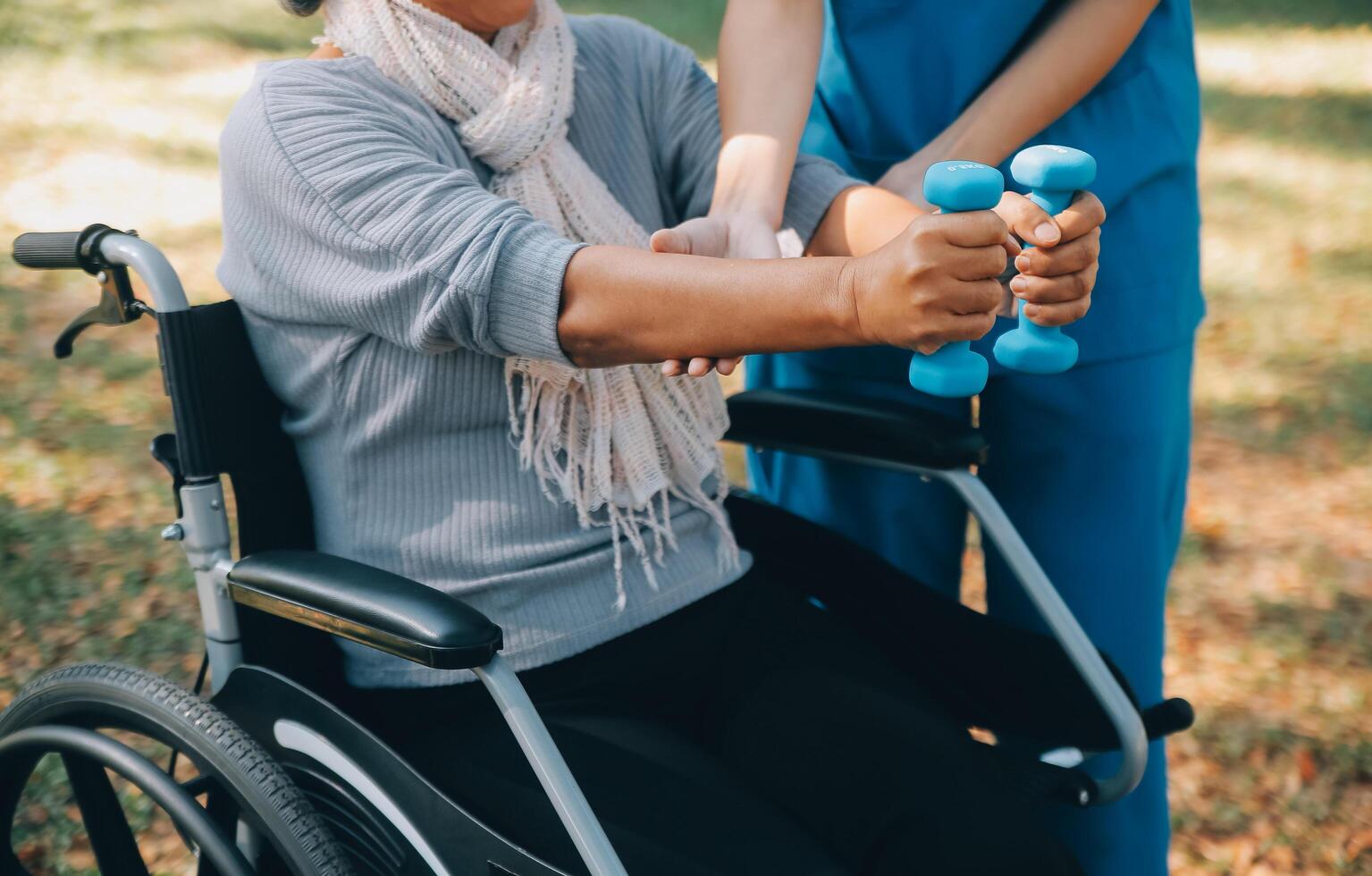 jong verpleegster of fysiotherapeut in scrubs helpen een gelukkig gepensioneerd oud vrouw Doen geschiktheid opdrachten met licht gewicht halters Bij huis. concept van fysiotherapie voor senioren foto