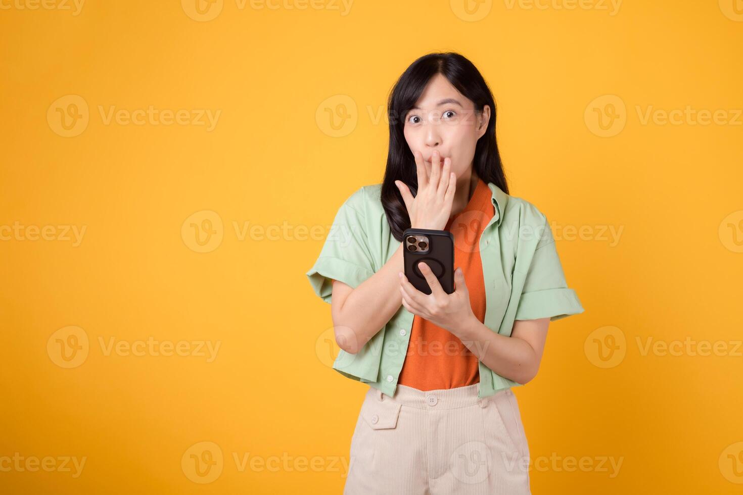 verrassing met een levendig jong Aziatisch vrouw in haar jaren '30, elegant gekleed in een oranje overhemd en groen trui, verdiept in haar smartphone Aan een geel studio achtergrond. app smartphone concept. foto