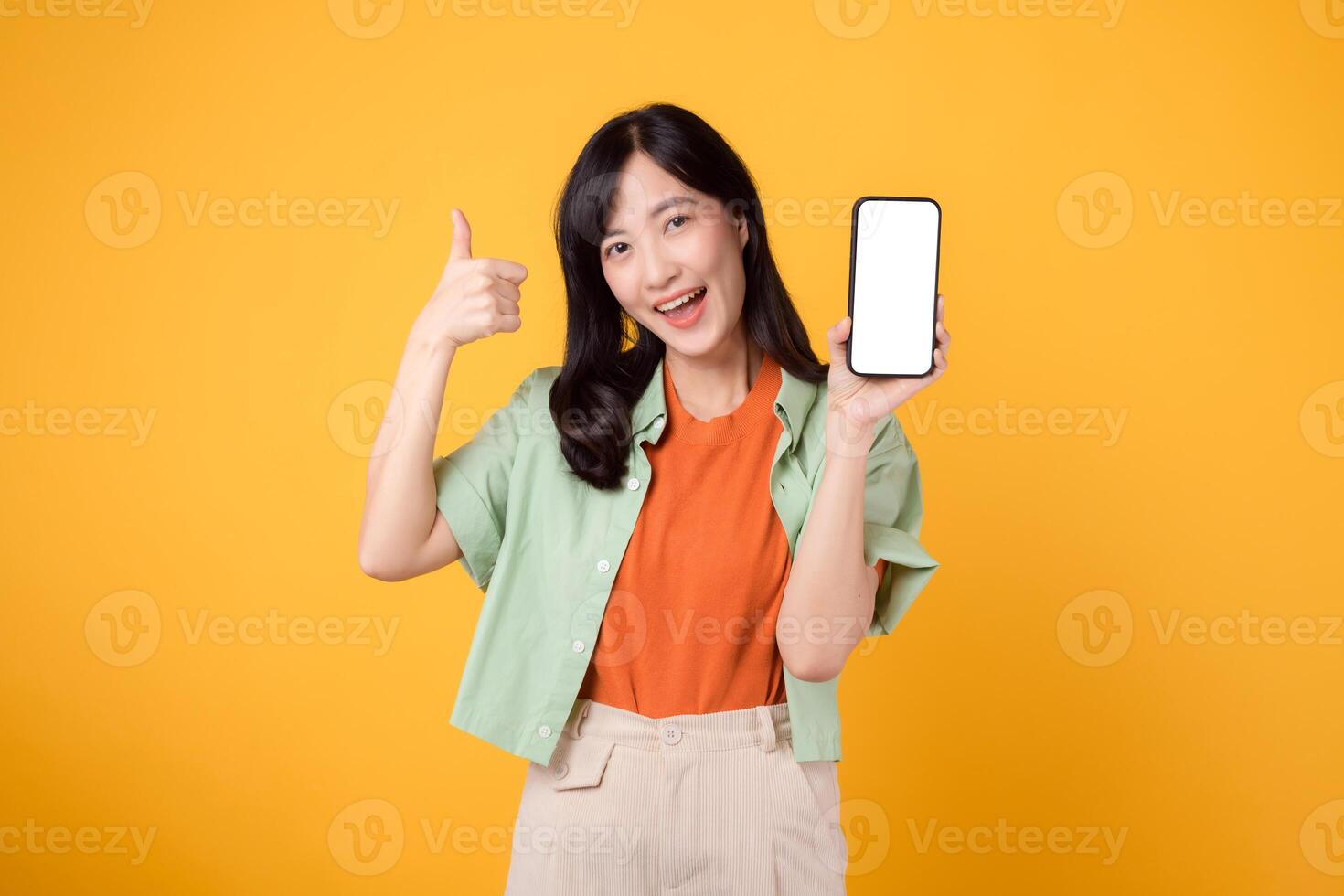 omhelzing de toekomst van nieuw mobiel toepassing met jong Aziatisch vrouw in haar jaren '30, vervelend oranje overhemd en groen trui, onthullend smartphone scherm met een duimen omhoog gebaar Aan geel studio achtergrond. foto