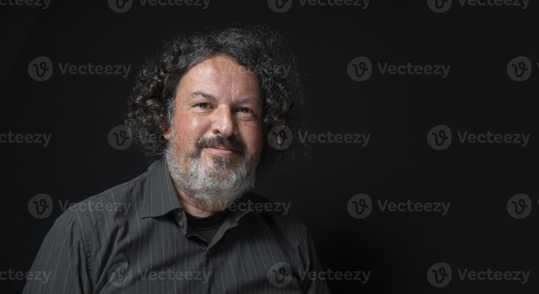 Mens met wit baard en zwart gekruld haar- met gelukkig en kalmte uitdrukking, op zoek naar camera, vervelend zwart overhemd tegen zwart achtergrond foto