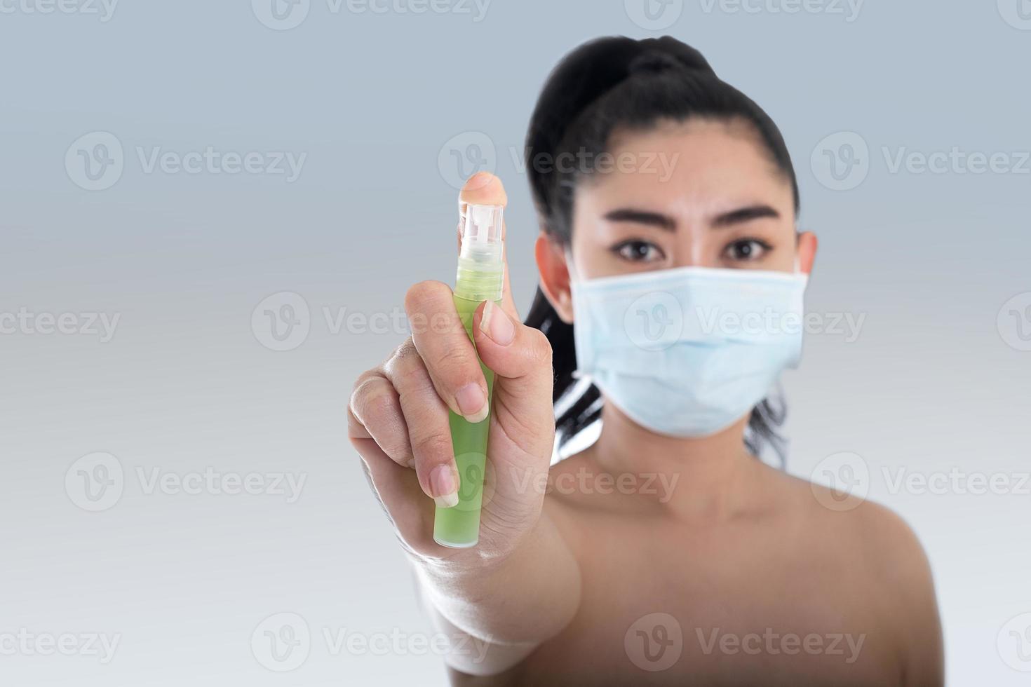 jonge aziatische vrouw die een medisch masker n95 opzet met de hand die alcoholspray uit de plastic fles of antibacteriën aanbrengt om de verspreiding van ziektekiemen op een grijze achtergrond te voorkomen foto