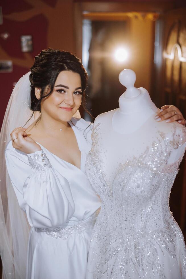 een gelukkig bruid is voorbereidingen treffen voor haar luxueus bruiloft in een hotel kamer, met een bruiloft jurk Aan een mannequin in de buurt. portret van een vrouw met modieus haar, bedenken en een glimlach in een dressing gewaad. foto