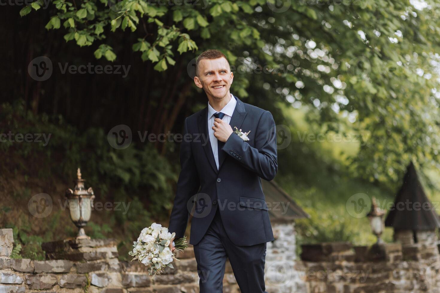 de bruidegom in een zwart pak past zich aan zijn boog binden, poses tegen de achtergrond van een groen boom. bruiloft portret. foto