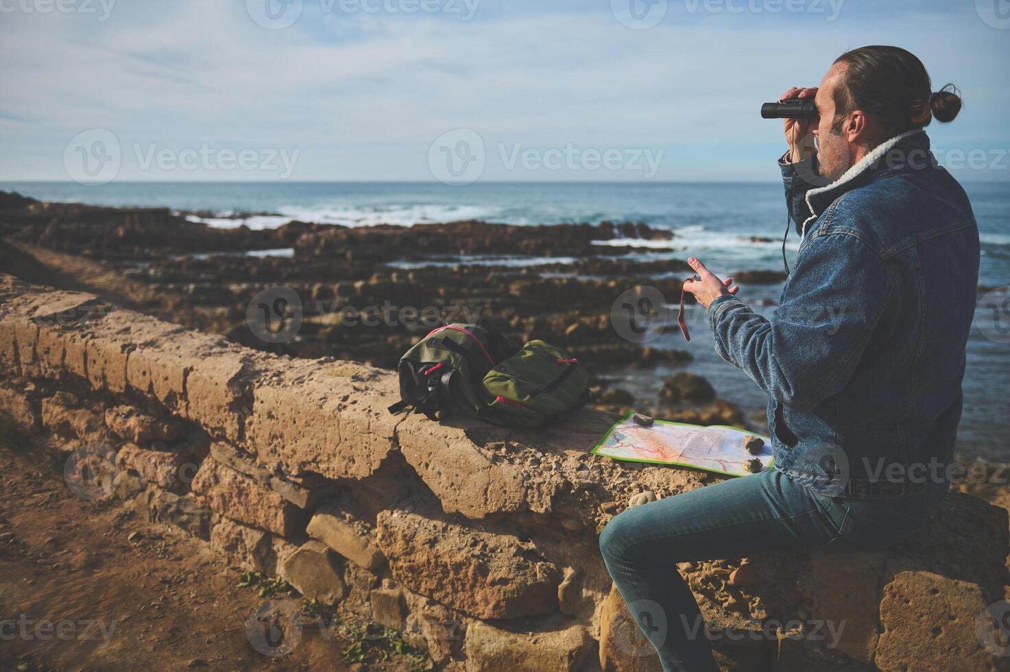 jong avonturier Holding kompas en gebruik makend van verrekijker, op zoek door naar de afstand, zittend Aan de rots door oceaan foto