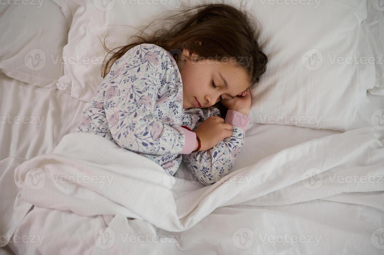 top visie mooi lief weinig kind meisje in elegant pyjama, slapen Aan haar bed, gedekt met wit schoon lakens foto