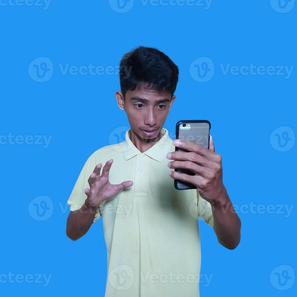 jong Aziatisch Mens verrast op zoek Bij slim telefoon vervelend geel t-shirt, blauw achtergrond. foto