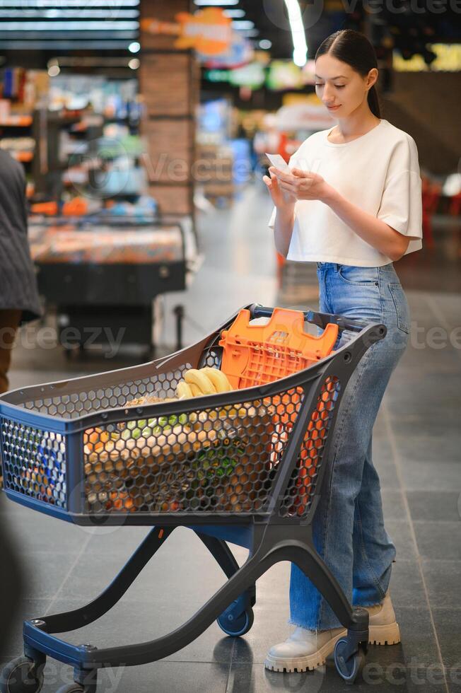 gelukkig jong vrouw op zoek Bij Product Bij kruidenier op te slaan. glimlachen vrouw boodschappen doen in supermarkt foto