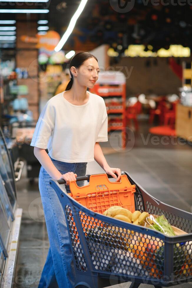 glimlachen gelukkig vrouw genieten van boodschappen doen Bij de supermarkt, ze is leunend Aan een vol kar foto