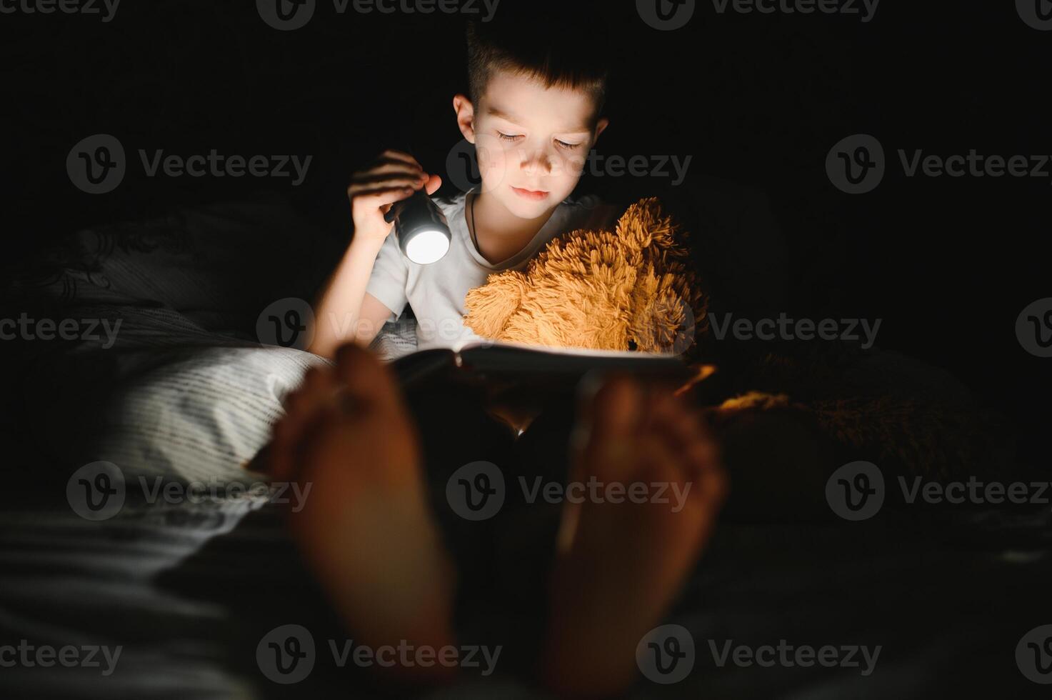 een jongen van 5-6 jaren oud is lezing een boek in de avond in de donker onder een deken met een speelgoed- beer. foto