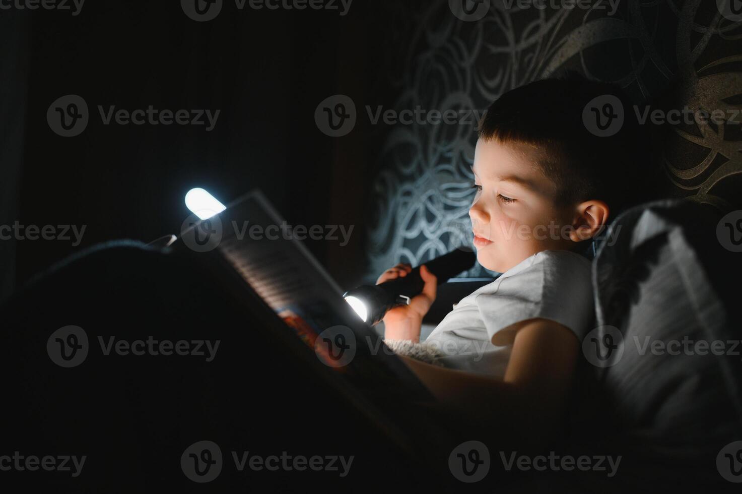 kind lezing boek in bed. kinderen lezen Bij nacht. weinig jongen met fee verhaal boeken in slaapkamer . onderwijs voor jong kinderen. bedtijd verhaal in de avond. schattig kind onder deken in donker kamer met lamp foto