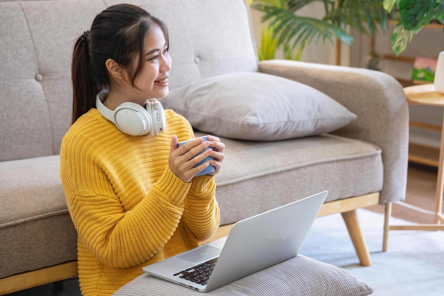 mooi jong vrouw in een geel gewoontjes jurk genieten van luisteren naar muziek- en glimlachen terwijl ontspannende Aan de sofa Bij huis. jong vrouw met hoofdtelefoons toepassingen laptop en smartphone Bij huis. kom tot rust concept foto