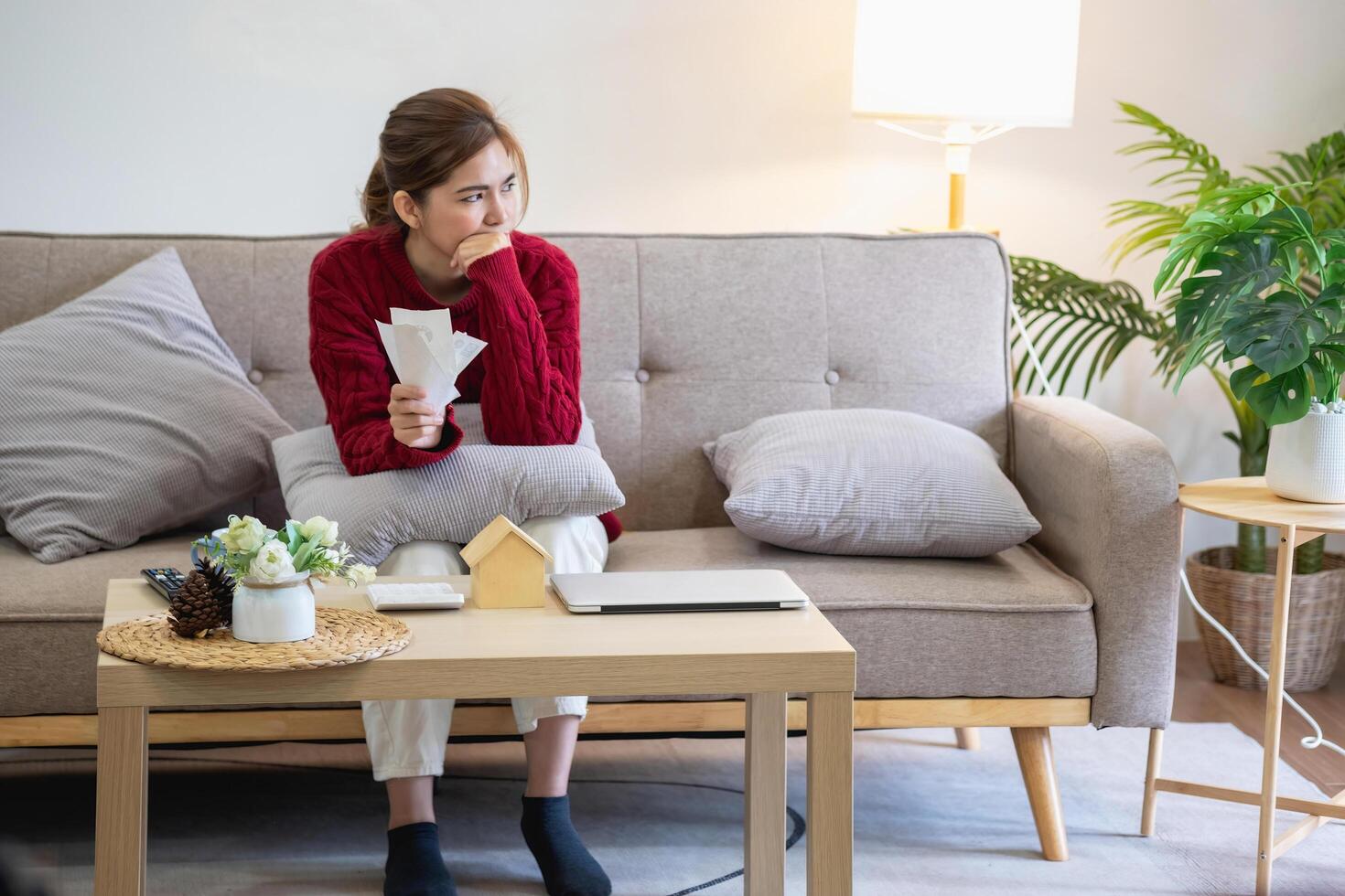 een jong Aziatisch vrouw zit Aan een sofa in haar huis, gevoel bezorgd en gefrustreerd over haar maandelijks uitgaven. divers nut rekeningen. foto