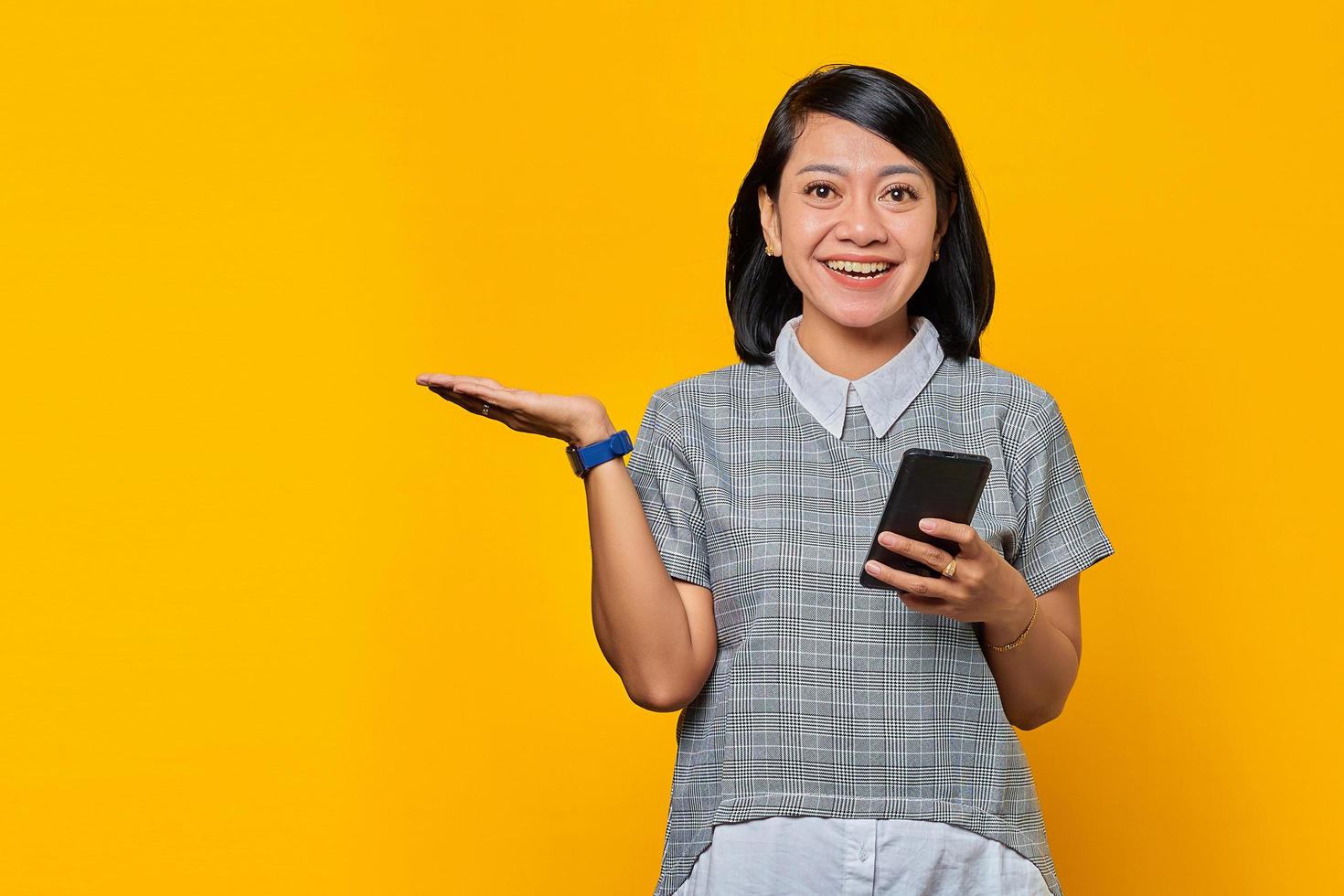 portret van een glimlachende jonge vrouw die kopieerruimte in de handpalm laat zien en een mobiele telefoon op een gele achtergrond houdt foto