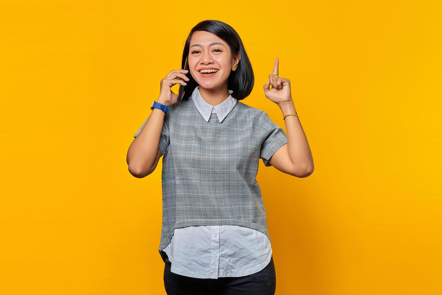 portret van opgewonden jonge aziatische vrouw die smartphone vasthoudt met een idee of vraag die met de vinger op gele achtergrond wijst foto