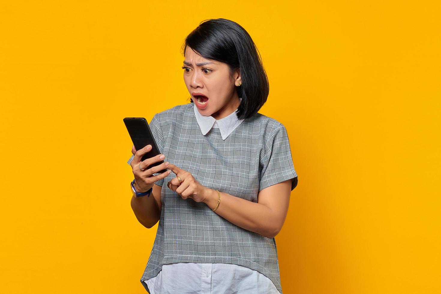 verraste jonge aziatische vrouw die een mobiele telefoon gebruikt en het smartphonescherm op gele achtergrond kijkt foto