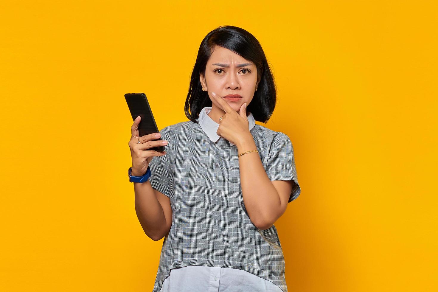 ongelukkige jonge aziatische vrouw met vinger op kin en mobiele telefoon vasthoudend over gele achtergrond foto