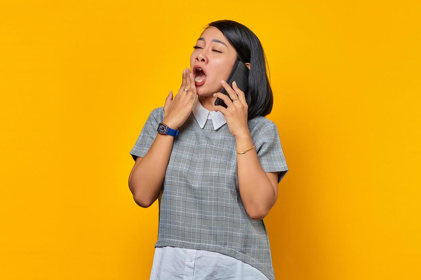 jonge aziatische vrouw geeuwt moe haar mond met de hand te bedekken terwijl ze een inkomende oproep ontvangt op een smartphone op een gele achtergrond foto