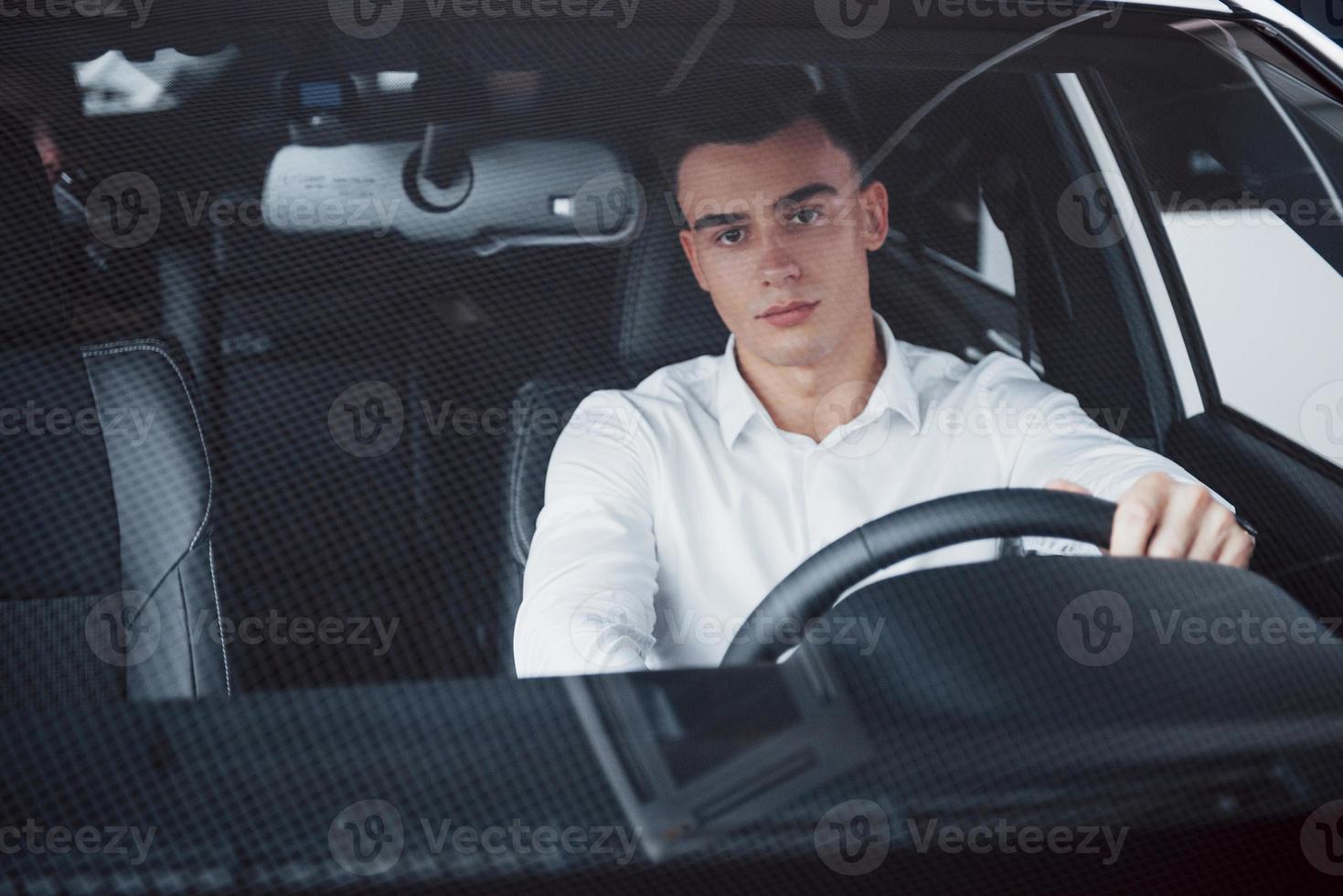 een jonge man zit in een pas gekochte auto en houdt zijn handen aan het roer foto