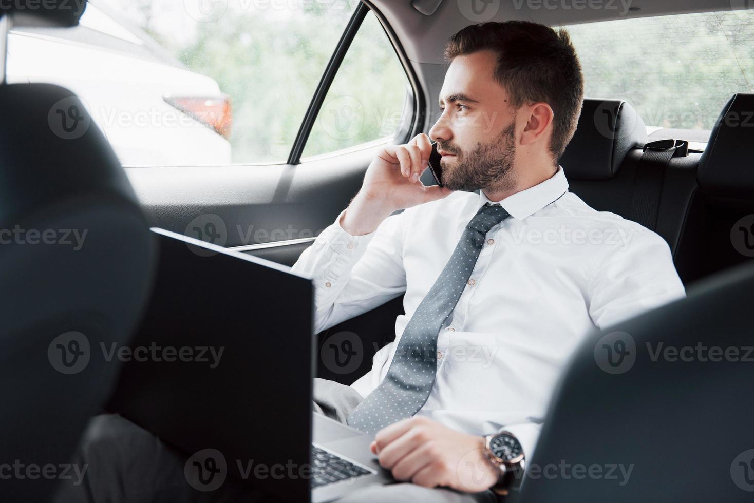 een jonge zakenman die aan een laptop werkt en aan de telefoon praat terwijl hij in de auto zit. werkt in beweging, waardeert zijn tijd foto