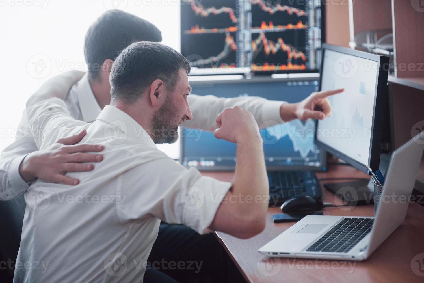 zakenlieden die online aandelen verhandelen. effectenmakelaars kijken naar grafieken, indexen en cijfers op meerdere computerschermen. collega's in discussie in het kantoor van de handelaar. zakelijk succes concept foto