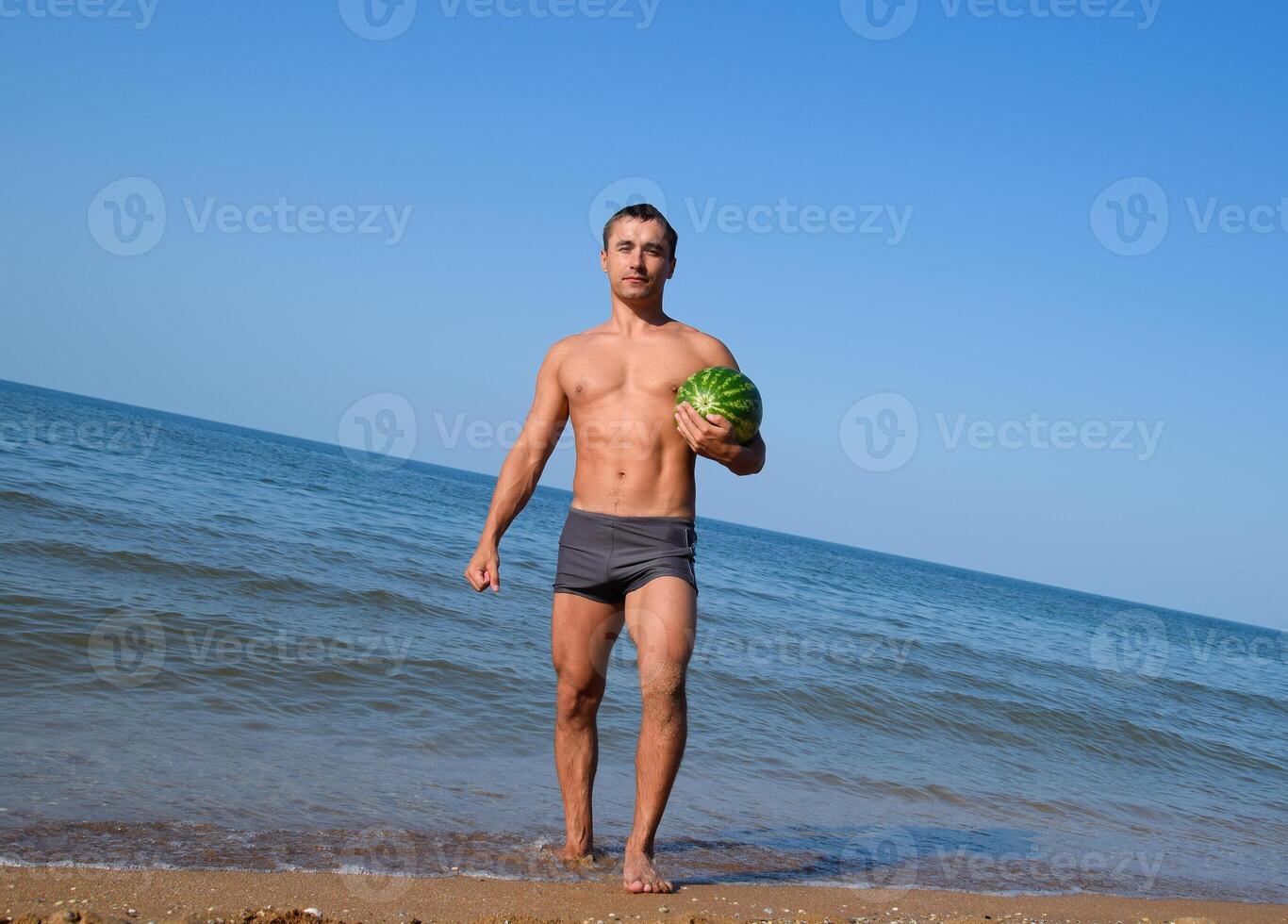 een sportief Mens is staand Aan de strand en Holding een geheel watermeloen. een rijp watermeloen in de handen van een Mens foto