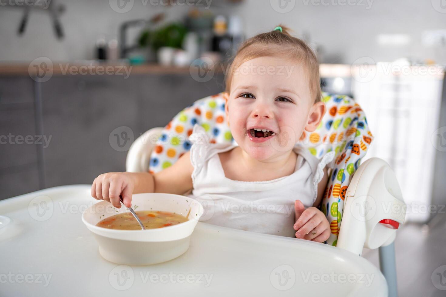 schattig baby meisje kleuter zittend in de hoog stoel en aan het eten haar lunch soep Bij huis keuken. foto