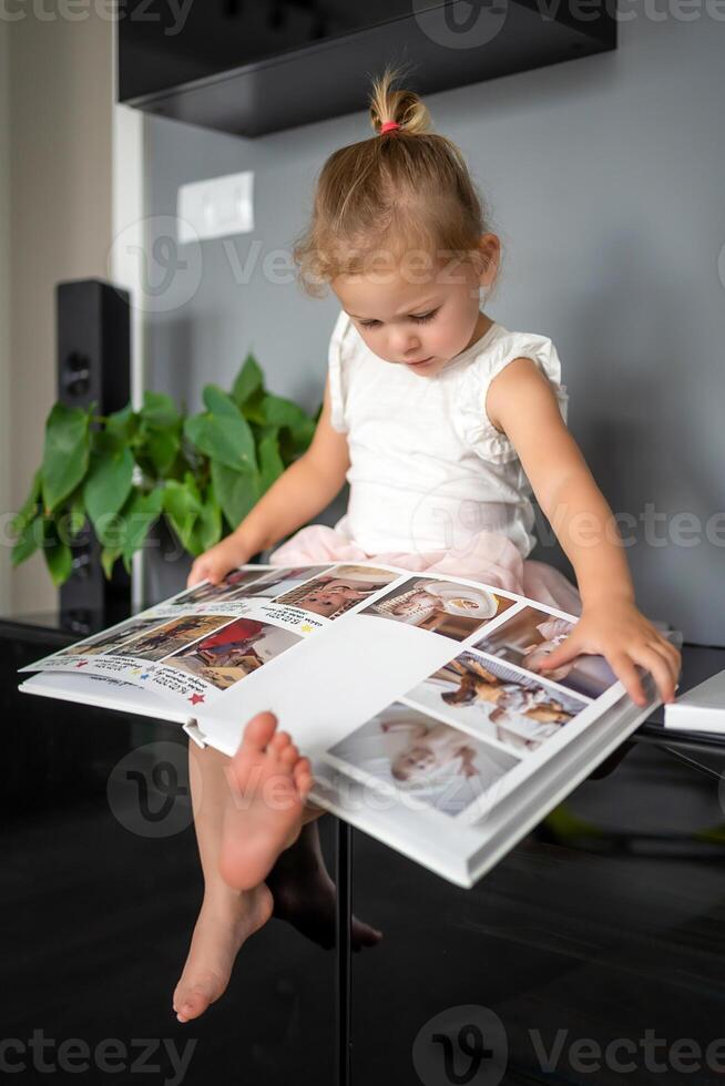gelukkig schattig weinig meisje op zoek Bij foto's in album. gelukkig glimlachen dochter browsen door gedrukt familie album, op zoek Bij afbeeldingen. foto