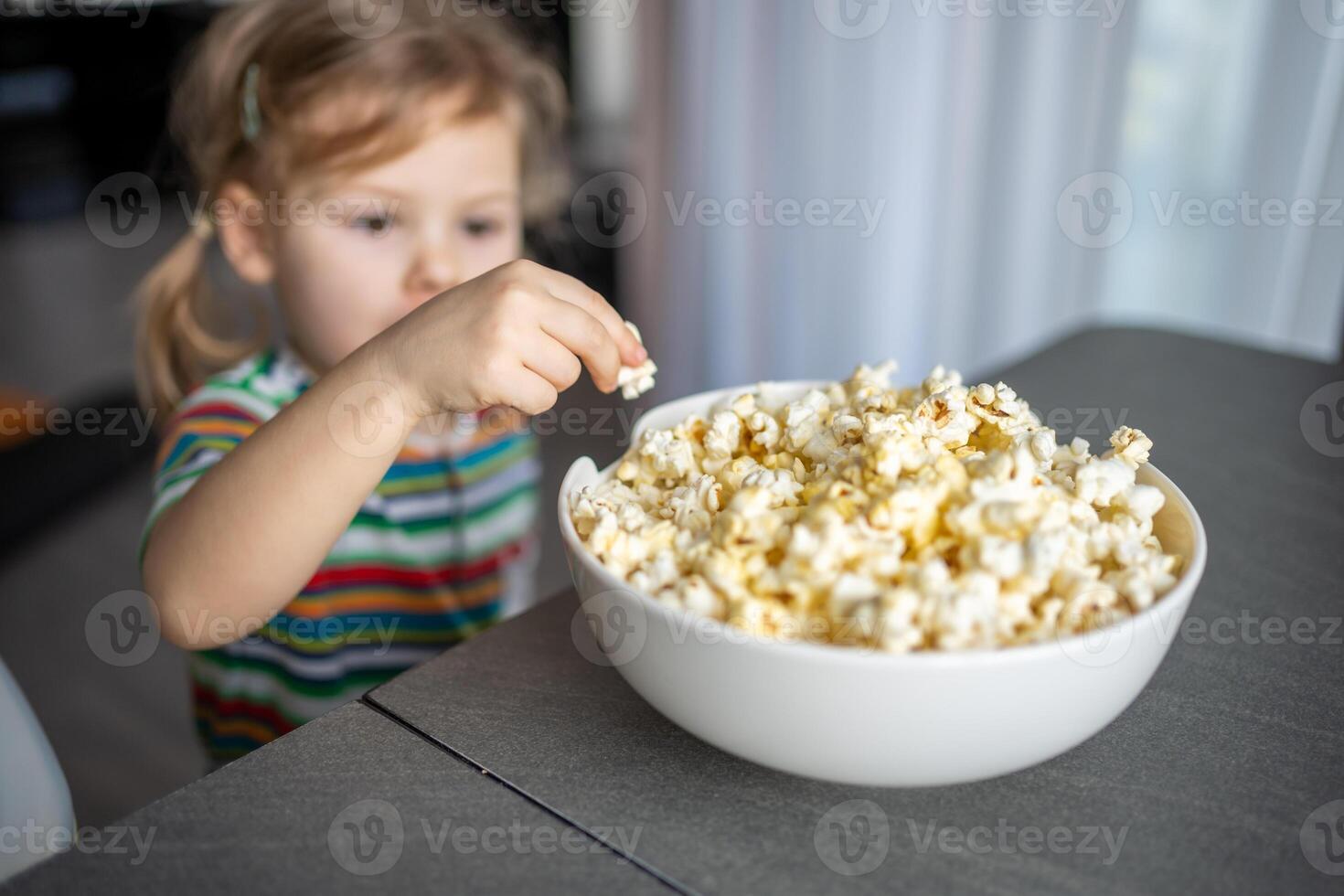 weinig meisje is aan het eten popcorn in huis keuken. focus Aan hand- nemen popcorn foto