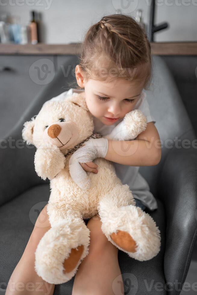 weinig meisje met gebroken vinger houdt teddy beer met een verbonden poot Bij de dokter afspraak in de ziekenhuis foto