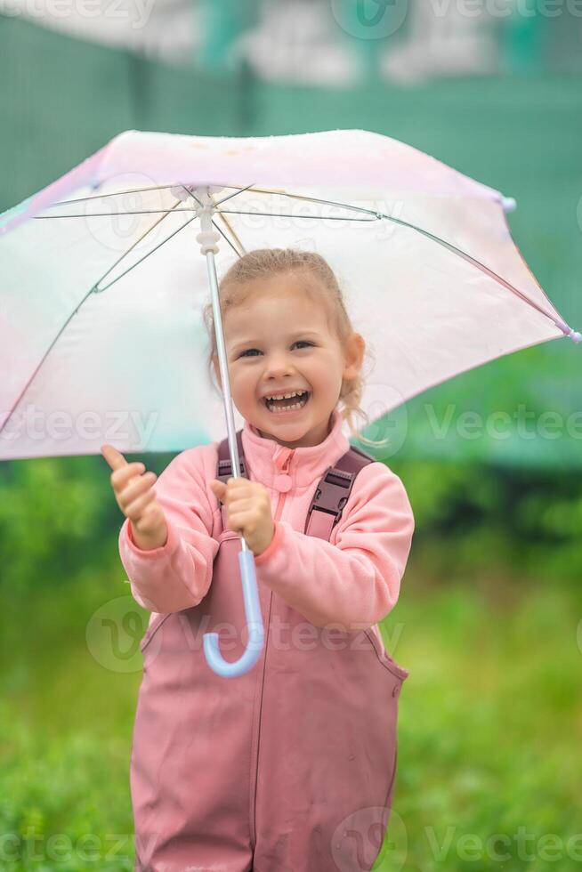 gelukkig lachend kind meisje 2-3 jaar oud vervelend waterbestendig kleren en Holding roze paraplu hebben een pret Aan huis achtertuin in regenachtig dag foto