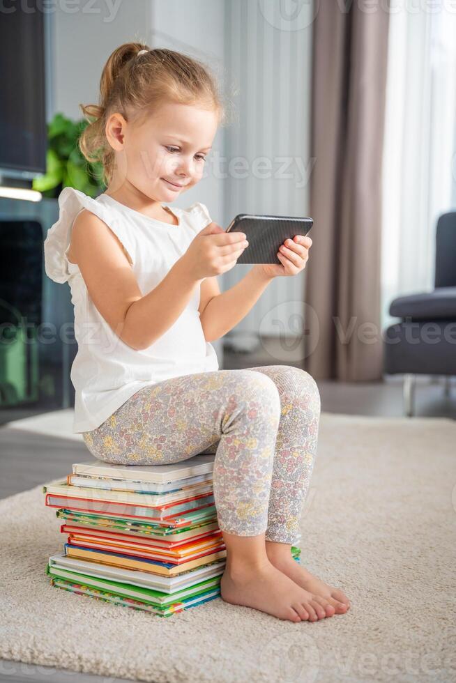 glimlachen weinig meisje zit Aan stack van kinderen sprookje boeken en horloges tekenfilms Aan smartphone foto