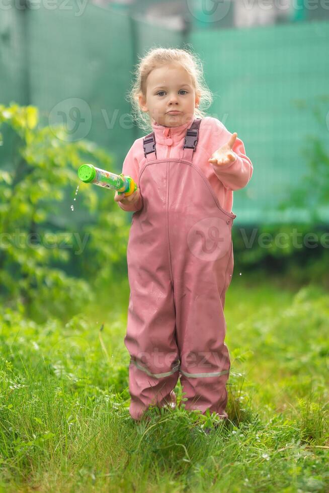 gelukkig lachend kind meisje 2-3 jaar oud vervelend waterbestendig kleren hebben een pret met water geweer Aan huis achtertuin foto