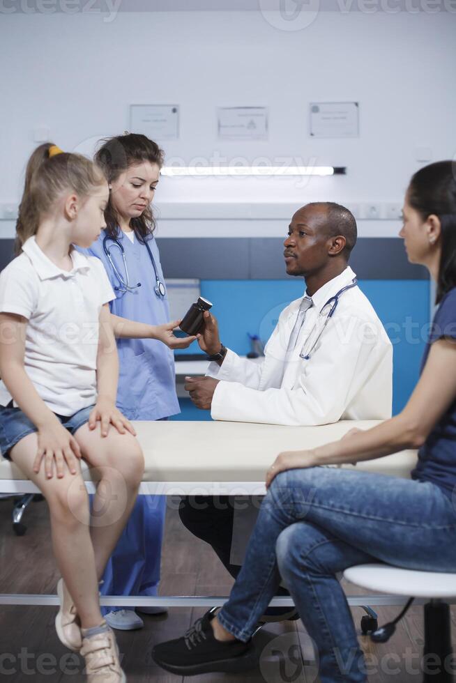 dokter geeft voorschrift pillen fles naar bijstand verpleegster in medisch kantoor. Kaukasisch moeder met jong meisje Bij gezondheidszorg kantoor ontvangen behandeling van kinderarts, krijgen advies Aan medicatie. foto