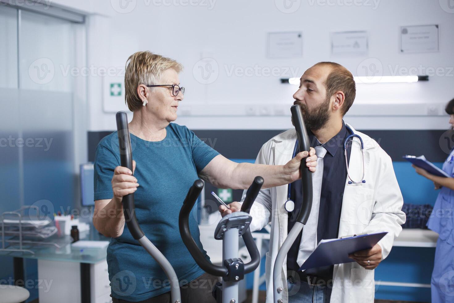 terwijl mannetje arts assists met fysiotherapie, ouderen geduldig bezighoudt in fysiek oefening Aan stationair fiets. naar herstellen van blessure, gepensioneerd vrouw ritten elektrisch fiets voor oefening. foto