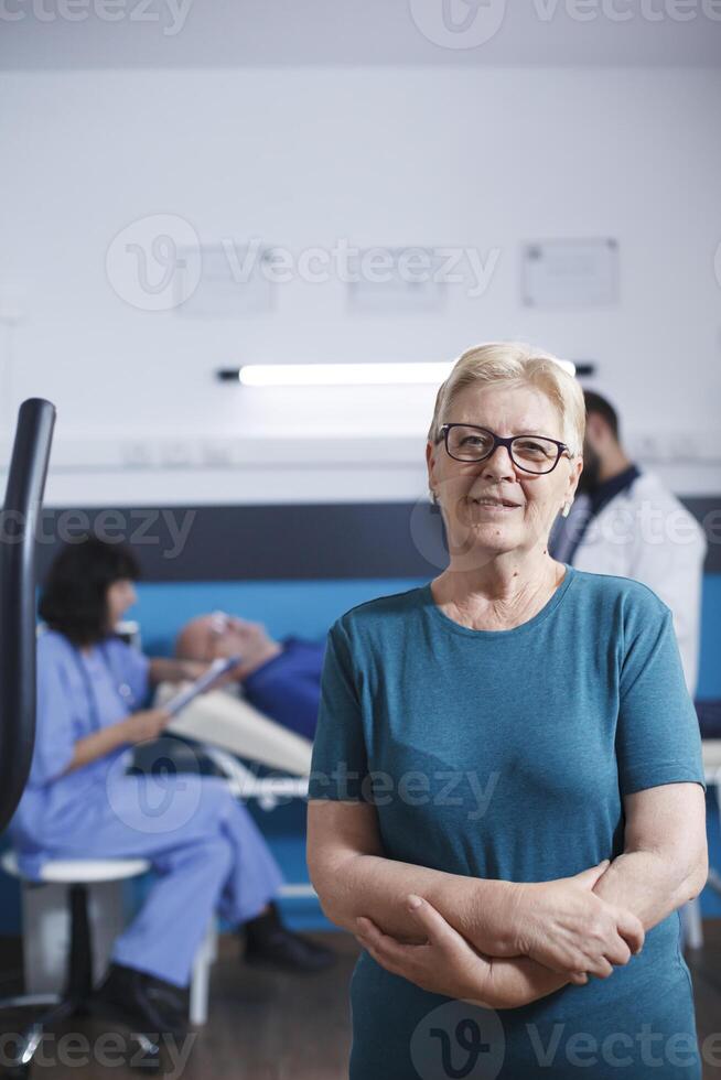 oud vrouw in portret wacht naar werk uit voor fysiek behandeling met armen gekruist. senior geduldig met gespierd letsel krijgt klaar voor been fysiotherapie Aan stationair fiets terwijl ze looks Bij camera. foto