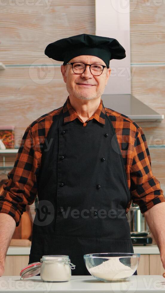 portret van chef vervelend bot op zoek Bij camera en lachend. gepensioneerd ouderen bakker in keuken uniform voorbereidingen treffen gebakje ingrediënten Aan houten tafel klaar naar koken eigengemaakt smakelijk brood, cakes en pasta foto