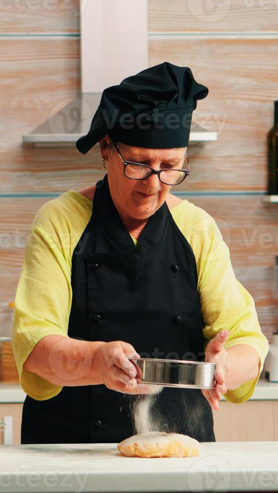 vrouw bakker gebruik makend van meel metalen zeef voorbereidingen treffen eigengemaakt taarten. gelukkig ouderen chef met bot voorbereidingen treffen rauw ingrediënten naar bakken traditioneel brood besprenkelen, zeven in de keuken. foto