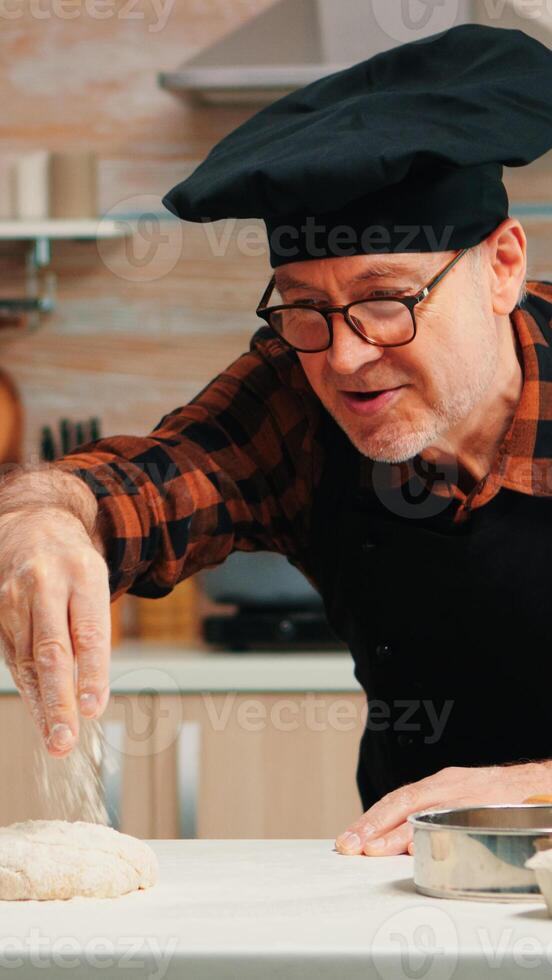 bakkerij Mens zeven meel over- deeg Aan tafel in huis keuken. gepensioneerd ouderen chef met bot en uniform besprenkelen, zeven, verspreiden rew ingrediënten met hand- bakken eigengemaakt pizza en brood. foto