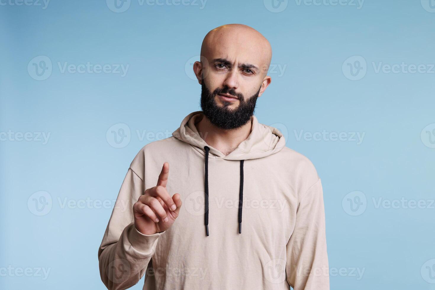 echt Arabisch Mens richten Bij camera en tonen een studio portret. jong volwassen kaal gebaard Arabisch persoon verhogen inhoudsopgave vinger en op zoek met zelfverzekerd gelaats uitdrukking foto