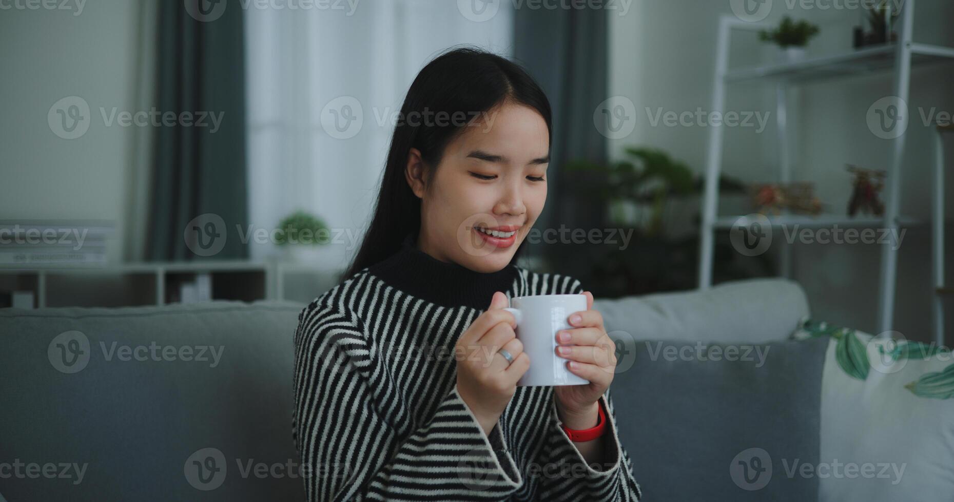 portret van ontspannende jong vrouw Holding kop genieten geur koffie of thee en drinken met geluk in ochtend- terwijl zitten Aan sofa in leven kamer, gratis tijd, nemen breken in thuis, glimlachend foto