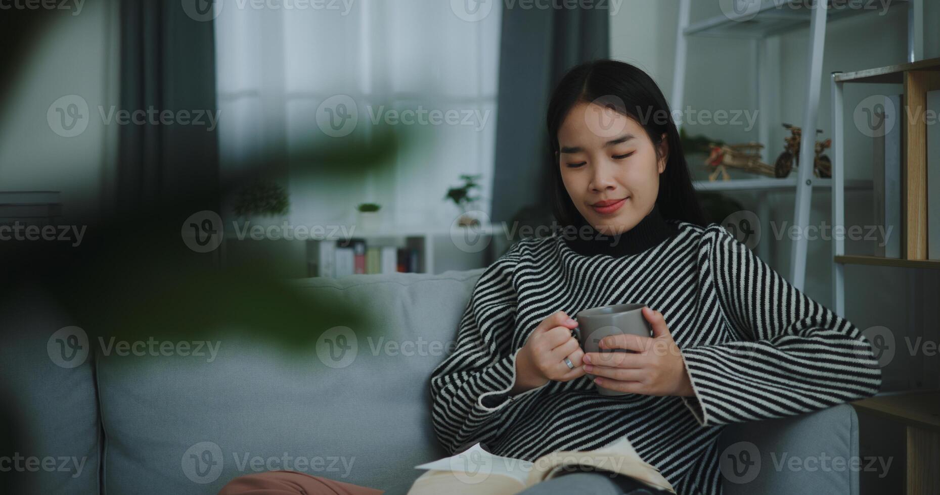 portret van gelukkig jong Aziatisch vrouw drinken ochtend- koffie of thee en lezing in leven kamer Bij huis Aan weekend. vrije tijd en levensstijl, gratis tijd foto