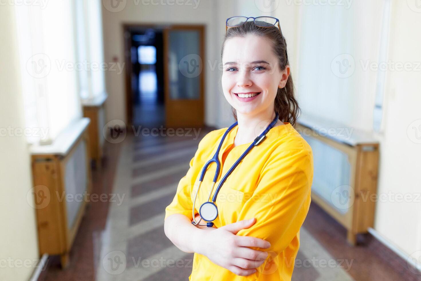 blij jong verpleegster. glimlachen jong meisje in geel uniform, en op zoek Bij camera, in voorkant van venster kliniek. de concept van Gezondheid foto