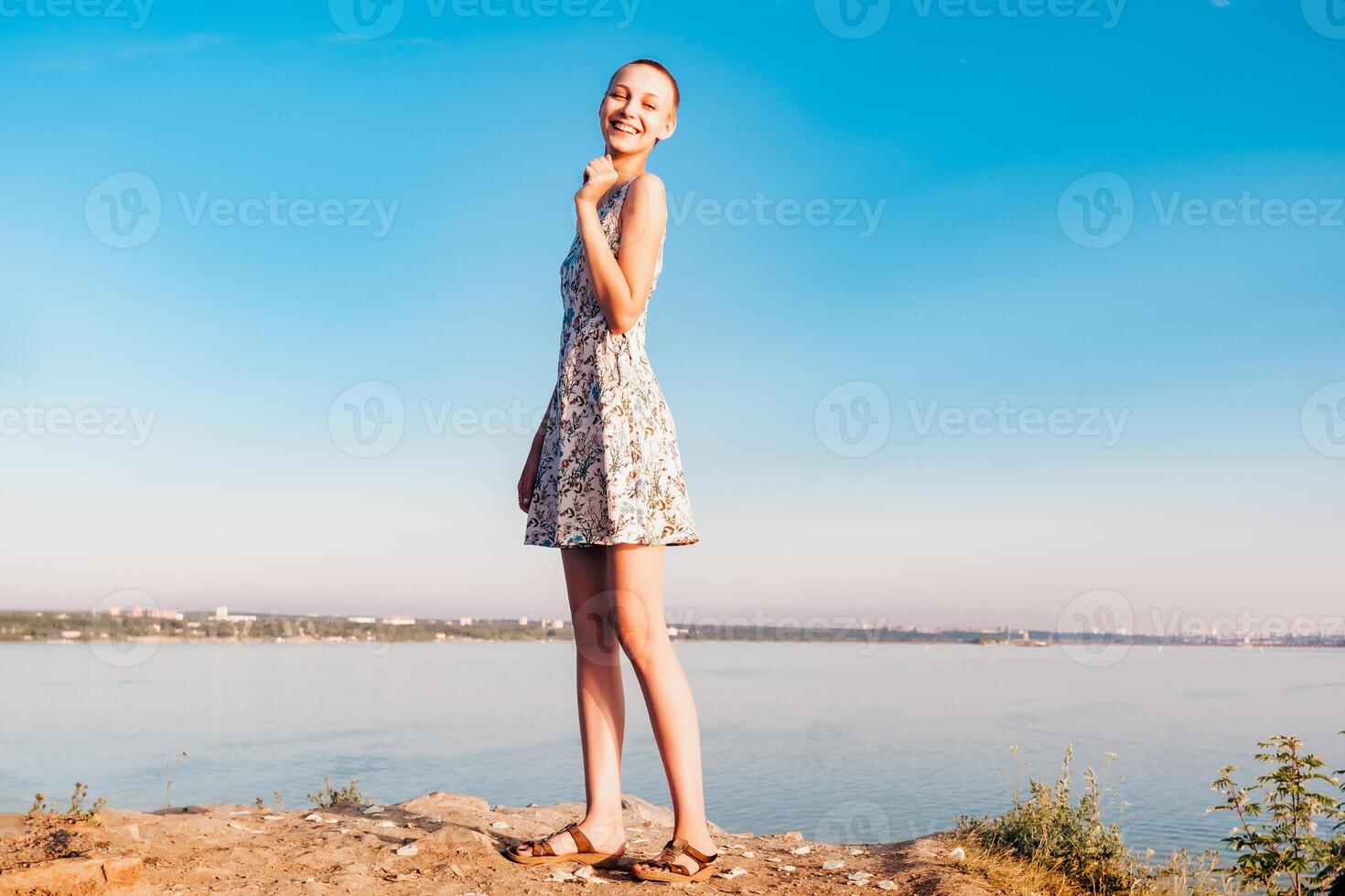lachend tiener meisje in een kort haren jurk staat Aan een klif over- een breed rivier- foto