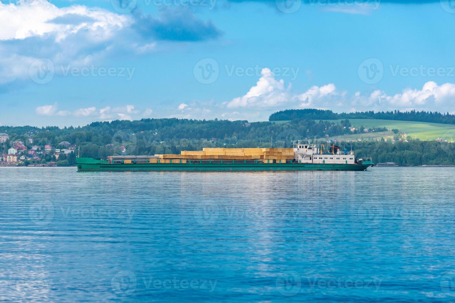 lading schip geladen met timmerhout volgt een breed rivier- Aan een zonnig dag foto